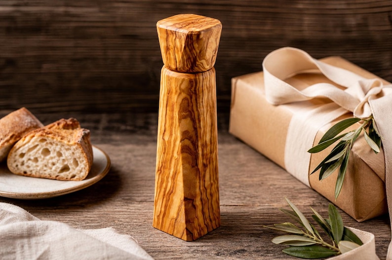 Pu&ograve; includere: Un macinapepe in legno dal design geometrico, che mostra la venatura naturale del legno. Nell'immagine sono presenti anche un piatto con fette di pane e un regalo avvolto con un nastro e del verde. Il macinapepe &egrave; alto circa 18 cm.