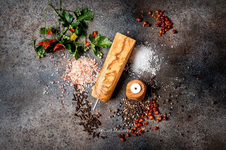 Pu&ograve; includere: Vista dall'alto di un macinapepe e di un saliera in legno con varie spezie ed erbe. La scena comprende sale rosa dell'Himalaya, grani di pepe nero, scaglie di peperoncino rosso e verde fresco, su una superficie strutturata grigio scuro.