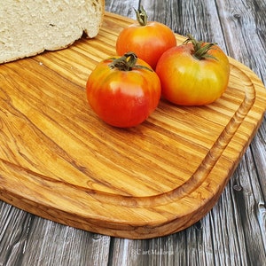 Op de afbeelding: Een houten snijplank met een afgeronde rand, gemaakt van olijfhout, met een brood en drie rode en gele tomaten erop.