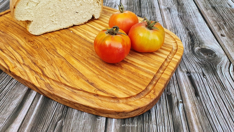 Op de afbeelding: Een houten snijplank met een afgeronde rand, gemaakt van olijfhout, met een brood en drie rode en gele tomaten erop.