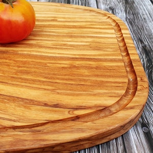 Op de afbeelding: Een groot, rechthoekig houten snijplank met een afgeronde rand en een natuurlijke houtnerf. De plank is gemaakt van olijfhout en heeft een lichtbruine kleur. Drie rode tomaten liggen op de plank.