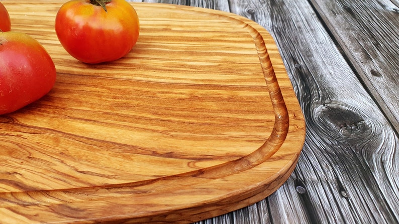 Op de afbeelding: Een groot, rechthoekig houten snijplank met een afgeronde rand en een natuurlijke houtnerf. De plank is gemaakt van olijfhout en heeft een lichtbruine kleur. Drie rode tomaten liggen op de plank.