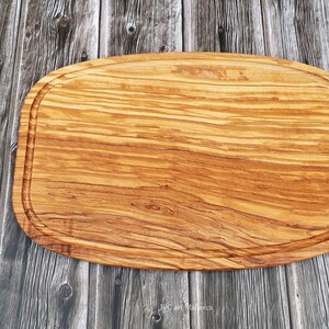 Op de afbeelding: Een groot, ovaal houten snijplank met een natuurlijke houtnerf afwerking. De plank heeft een verhoogde rand en is gemaakt van olijfhout. Drie rode tomaten zijn in een rij links van de plank gerangschikt.