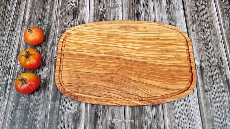 Op de afbeelding: Een groot, ovaal houten snijplank met een natuurlijke houtnerf afwerking. De plank heeft een verhoogde rand en is gemaakt van olijfhout. Drie rode tomaten zijn in een rij links van de plank gerangschikt.
