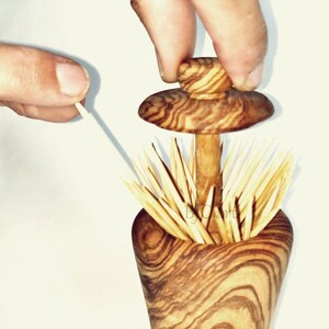 May include: A wooden toothpick holder shaped like a mushroom with a brown and white wood grain pattern. The top of the holder is a removable lid that can be lifted to access the toothpicks. The holder is filled with toothpicks.