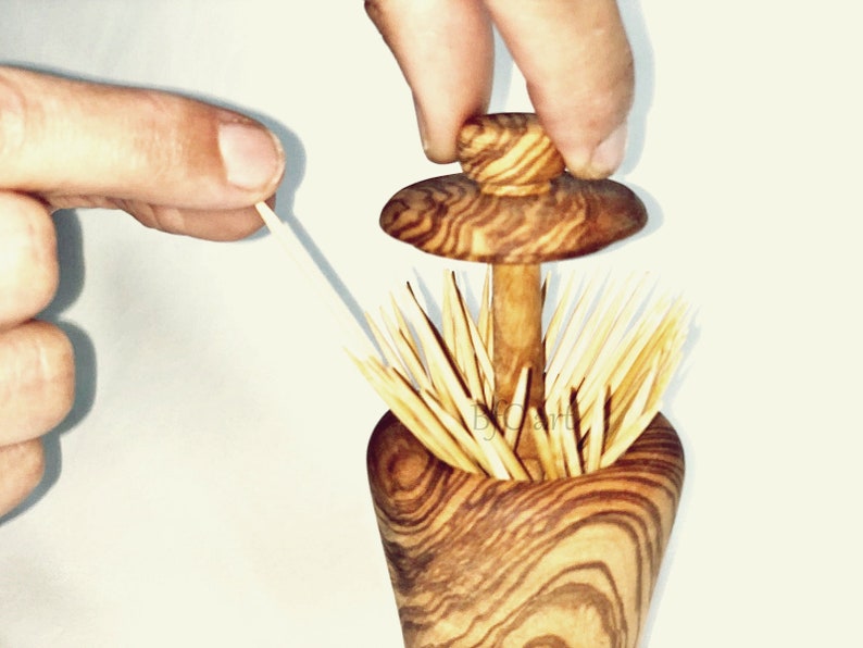 May include: A wooden toothpick holder shaped like a mushroom with a brown and white wood grain pattern. The top of the holder is a removable lid that can be lifted to access the toothpicks. The holder is filled with toothpicks.