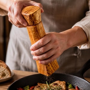 Pu&ograve; includere: Un macinapepe in legno &egrave; tenuto sopra una padella in ghisa. Il macinino &egrave; in legno marrone chiaro con una venatura strutturata. La padella contiene cibo cotto con verdure. Pane a fette &egrave; su un piatto sullo sfondo.