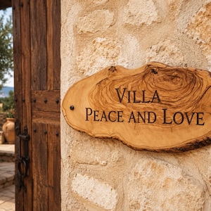 Puede incluir: Un letrero de madera con bordes naturales y una veta prominente, montado sobre una pared de piedra texturizada. El letrero presenta el texto grabado VILLA PEACE AND LOVE en una fuente serif oscura, asegurado con dos tornillos de metal oscuro.