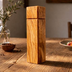 Op de afbeelding: Een hoge, rechthoekige houten pepermolen met een vierkante bovenkant en een natuurlijke houtnerf. De molen staat op een houten tafel, met een kom met peperkorrels en een salade op de achtergrond. Thema: keukengerei.