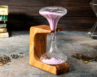 May include: A pink hourglass with a wooden stand. The hourglass is made of clear glass and has a bulbous shape. The sand is a light pink color. The stand is made of wood and has a natural finish.