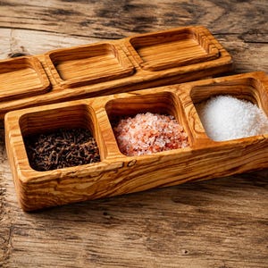 May include: Wooden spice serving dish with three compartments, one filled with cloves, one with pink salt, and one with white salt. The dish is made of light brown wood and sits on a wooden surface.