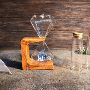 May include: A glass hourglass with a diamond-shaped design, resting on a wooden stand. Two small glass bottles with cork tops are also included.