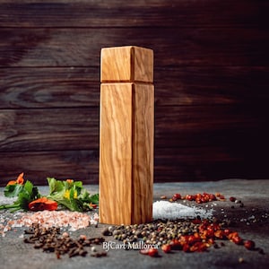 Pode incluir: Um moinho de sal e pimenta de madeira com formato quadrado e gr&atilde;o de madeira natural. O moinho est&aacute; sobre uma superf&iacute;cie cinza com v&aacute;rias especiarias e ervas espalhadas ao redor.