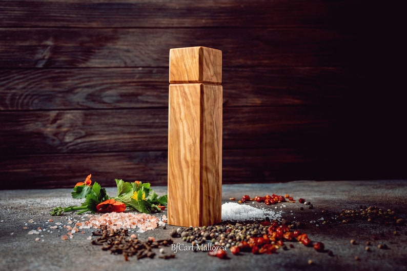 Pode incluir: Um moinho de sal e pimenta de madeira com formato quadrado e gr&atilde;o de madeira natural. O moinho est&aacute; sobre uma superf&iacute;cie cinza com v&aacute;rias especiarias e ervas espalhadas ao redor.