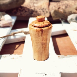 May include: A small, wooden jar with a lid, made of olive wood. The jar has a natural wood finish and is sitting on a white surface.