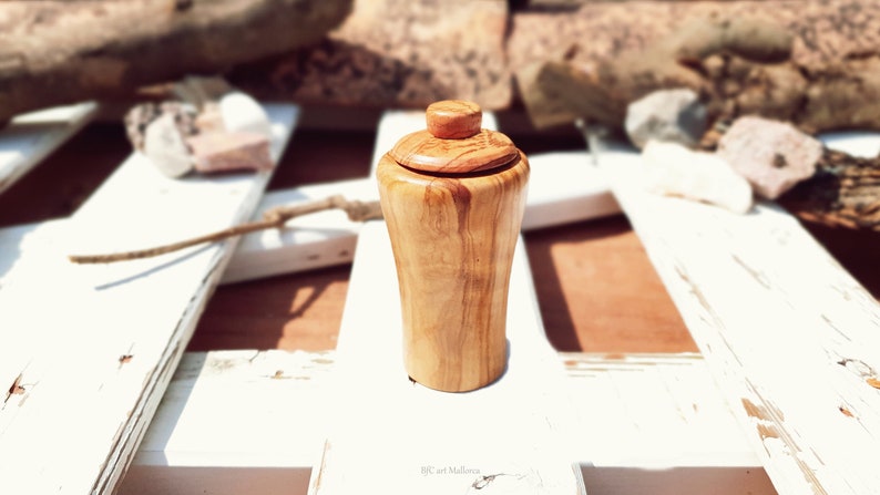 May include: A small, wooden jar with a lid, made of olive wood. The jar has a natural wood finish and is sitting on a white surface.