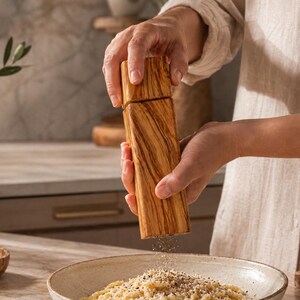 Op de afbeelding: Een houten pepermolen met een lichtbruine afwerking wordt boven een kom pasta gehouden. De molen wordt gebruikt om peper op de pasta te malen. De keuken omvat een marmeren aanrecht en olijfolie.