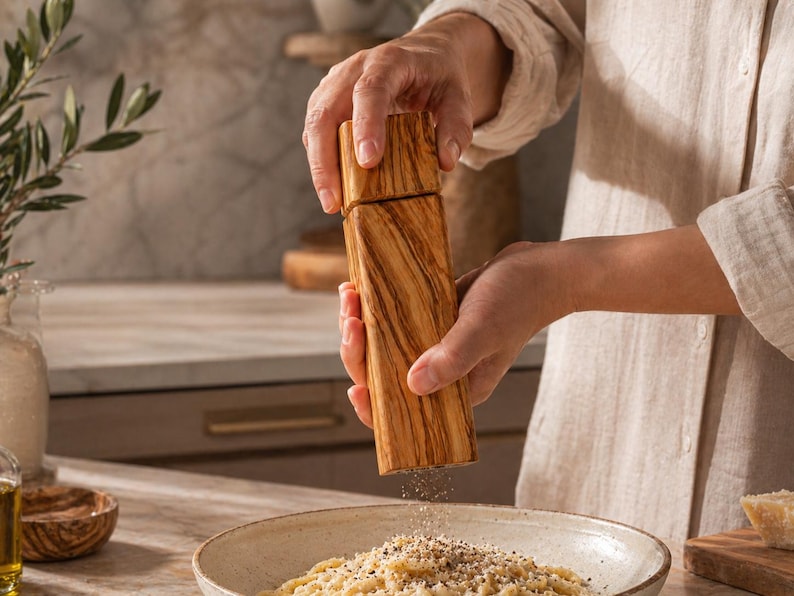 Op de afbeelding: Een houten pepermolen met een lichtbruine afwerking wordt boven een kom pasta gehouden. De molen wordt gebruikt om peper op de pasta te malen. De keuken omvat een marmeren aanrecht en olijfolie.