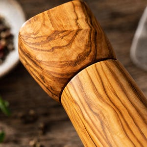 Pu&ograve; includere: Primo piano di un macinapepe in legno con venatura naturale. Il macinapepe ha un corpo cilindrico e una parte superiore sfaccettata. Il legno &egrave; di un marrone caldo con striature pi&ugrave; scure, forse legno d'ulivo. Lo sfondo sfocato mostra spezie.