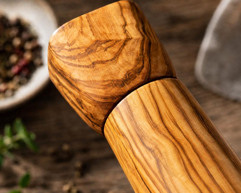 Pu&ograve; includere: Primo piano di un macinapepe in legno con venatura naturale. Il macinapepe ha un corpo cilindrico e una parte superiore sfaccettata. Il legno &egrave; di un marrone caldo con striature pi&ugrave; scure, forse legno d'ulivo. Lo sfondo sfocato mostra spezie.