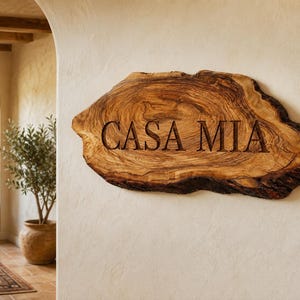 Puede incluir: Un letrero de madera natural con borde vivo y un patr&oacute;n de veta prominente, que presenta el texto "CASA MIA" grabado profundamente en una fuente serif oscura. La pieza tiene una forma org&aacute;nica irregular con bordes de corteza texturizados.
