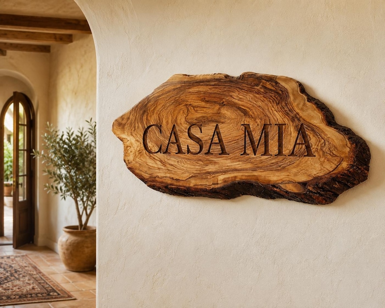 Puede incluir: Un letrero de madera natural con borde vivo y un patr&oacute;n de veta prominente, que presenta el texto "CASA MIA" grabado profundamente en una fuente serif oscura. La pieza tiene una forma org&aacute;nica irregular con bordes de corteza texturizados.