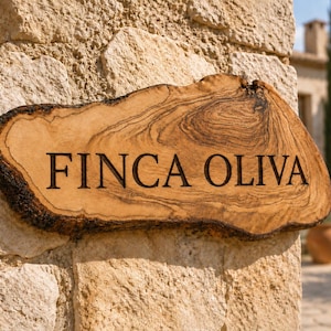 Può includere: Un'insegna in legno con bordo naturale che riporta il testo FINCA OLIVA inciso in lettere maiuscole nere. L'insegna presenta un marcato motivo a venature del legno con tonalità marrone chiaro e scuro, che ne esalta la forma e la consistenza.