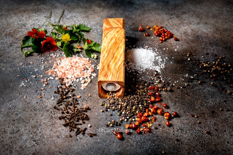 Pode incluir: Um moinho de pimenta de madeira com acabamento de gr&atilde;o de madeira natural est&aacute; sobre uma superf&iacute;cie cinza rodeado de v&aacute;rias especiarias, incluindo sal rosa do Himalaia, sal branco, gr&atilde;os de pimenta preta, gr&atilde;os de pimenta vermelha e cravos.