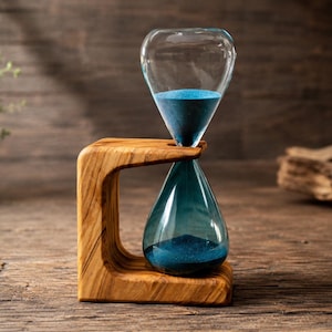 May include: An hourglass featuring a clear glass bulb filled with fine blue sand, held within a curved, light brown wooden frame with a distinct natural grain pattern. The structure supports the glass vessel in a vertical, suspended position.