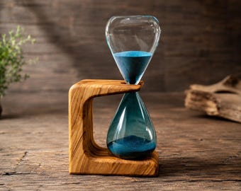 May include: An hourglass featuring a clear glass bulb filled with fine blue sand, held within a curved, light brown wooden frame with a distinct natural grain pattern. The structure supports the glass vessel in a vertical, suspended position.