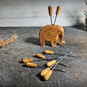 May include: A wooden elephant-shaped toothpick holder with two toothpicks and several metal and wood-handled picks. The holder is light brown, and the picks have light brown handles. The items are on a textured surface.