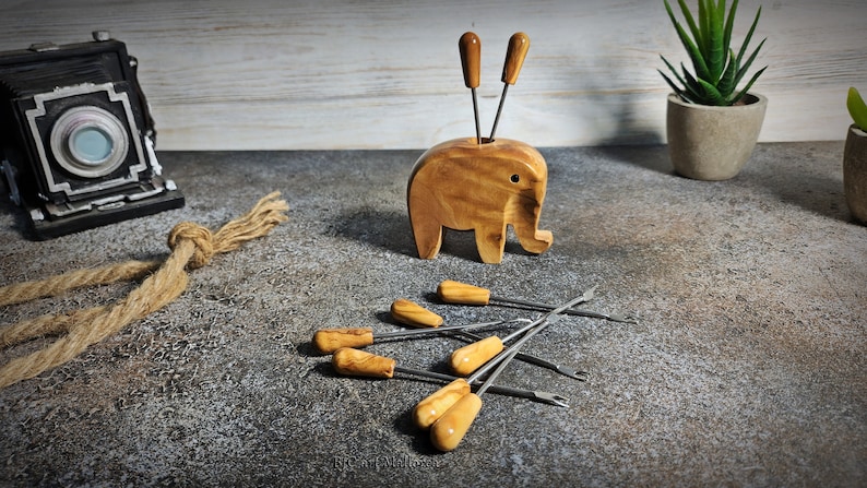May include: A wooden elephant-shaped toothpick holder with two toothpicks and several metal and wood-handled picks. The holder is light brown, and the picks have light brown handles. The items are on a textured surface.