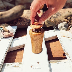 May include: A wooden toothpick holder with a lid. The holder is made of light brown wood with a natural grain pattern. The lid is a small, round piece of wood that fits snugly on top of the holder. The holder is sitting on a white wooden surface.