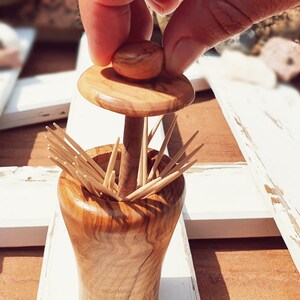 May include: A wooden toothpick holder with a lid. The holder is made of olive wood and has a natural finish. The lid is also made of olive wood and has a knob on top. The holder is filled with toothpicks.