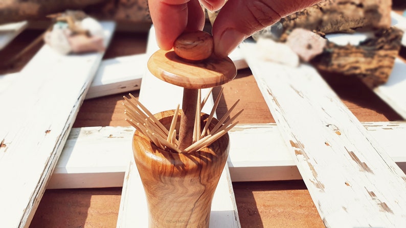 May include: A wooden toothpick holder with a lid. The holder is made of olive wood and has a natural finish. The lid is also made of olive wood and has a knob on top. The holder is filled with toothpicks.