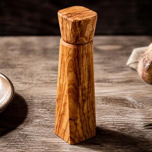 Pu&ograve; includere: Un macinapepe in legno dal design geometrico. Il macinapepe &egrave; realizzato in legno marrone chiaro con un motivo a venature naturali. Ha una parte superiore quadrata e un corpo affusolato. Una piccola ciotola di ceramica con sale e grani di pepe &egrave; sullo sfondo.