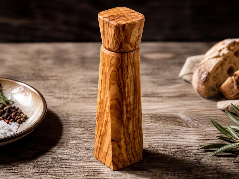 Pu&ograve; includere: Un macinapepe in legno dal design geometrico. Il macinapepe &egrave; realizzato in legno marrone chiaro con un motivo a venature naturali. Ha una parte superiore quadrata e un corpo affusolato. Una piccola ciotola di ceramica con sale e grani di pepe &egrave; sullo sfondo.
