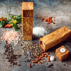 Pode incluir: Dois moinhos de sal e pimenta de madeira com topo quadrado e corpo retangular. Os moinhos s&atilde;o feitos de madeira de oliveira e t&ecirc;m uma veia de madeira natural. Os moinhos s&atilde;o rodeados por v&aacute;rias especiarias, incluindo sal rosa do Himalaia, sal branco, gr&atilde;os de pimenta preta, cravos e flocos de pimenta vermelha.