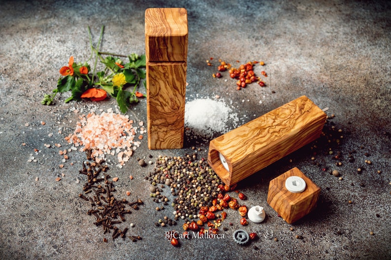 Pode incluir: Dois moinhos de sal e pimenta de madeira com topo quadrado e corpo retangular. Os moinhos s&atilde;o feitos de madeira de oliveira e t&ecirc;m uma veia de madeira natural. Os moinhos s&atilde;o rodeados por v&aacute;rias especiarias, incluindo sal rosa do Himalaia, sal branco, gr&atilde;os de pimenta preta, cravos e flocos de pimenta vermelha.