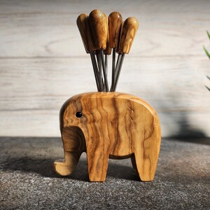 May include: A wooden elephant-shaped appetizer pick holder with six picks. The holder is made of light brown wood with a visible grain pattern. The picks have wooden handles and metal prongs. A small potted plant is in the background.