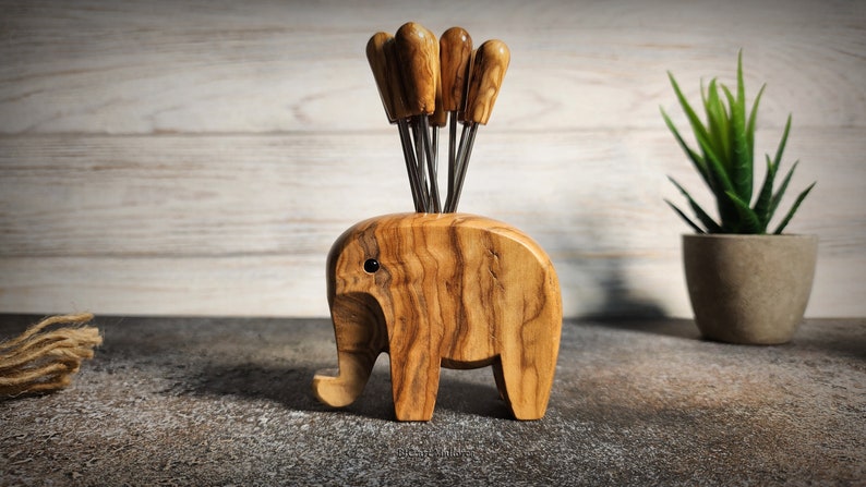 May include: A wooden elephant-shaped appetizer pick holder with six picks. The holder is made of light brown wood with a visible grain pattern. The picks have wooden handles and metal prongs. A small potted plant is in the background.