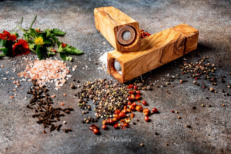 Pode incluir: Dois moinhos de sal e pimenta de madeira com acabamento em madeira natural. Os moinhos s&atilde;o rodeados por v&aacute;rias especiarias, incluindo sal rosa do Himalaia, gr&atilde;os de pimenta preta, gr&atilde;os de pimenta branca, flocos de pimenta vermelha e cravos.