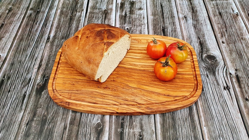 Op de afbeelding: Een houten snijplank met een lichtbruine afwerking en een licht ovale vorm. De plank heeft een verhoogde rand en een groef rondom de omtrek. Een brood en vier rode tomaten liggen op de plank.