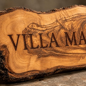 Puede incluir: Una placa de madera de borde natural con corteza en los extremos y una superficie de veta marr&oacute;n clara. El texto VILLA MAR est&aacute; grabado en una fuente serif oscura en el centro de la madera, resaltando su textura org&aacute;nica.