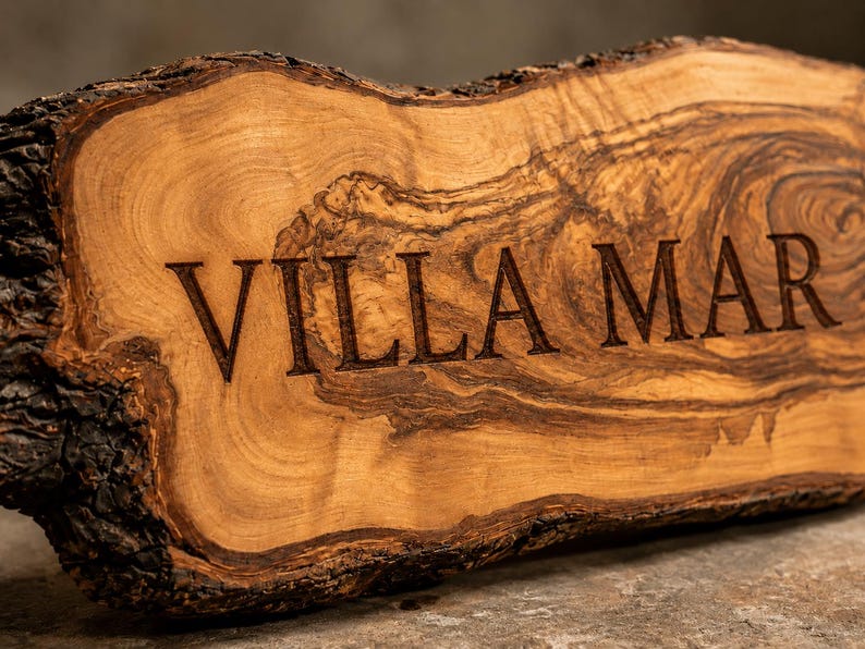 Puede incluir: Una placa de madera de borde natural con corteza en los extremos y una superficie de veta marr&oacute;n clara. El texto VILLA MAR est&aacute; grabado en una fuente serif oscura en el centro de la madera, resaltando su textura org&aacute;nica.