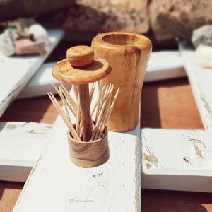 May include: Two wooden toothpick holders, one with a lid and the other with a round top, are displayed on a white wooden surface. The holders are made of olive wood and have a natural wood finish.
