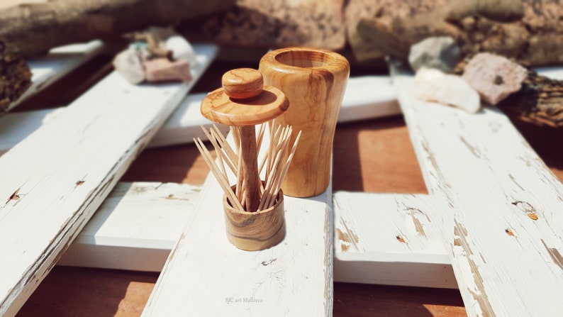 May include: Two wooden toothpick holders, one with a lid and the other with a round top, are displayed on a white wooden surface. The holders are made of olive wood and have a natural wood finish.