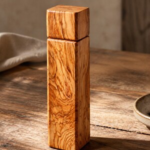 Op de afbeelding: Een rechthoekige houten pepermolen met een vierkante bovenkant en een natuurlijke houtnerf. De molen staat op een houten oppervlak, met een kleine kom met peperkorrels in de buurt. De algehele esthetiek is rustiek en elegant.