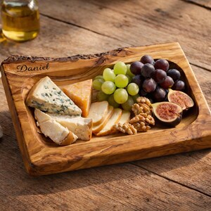 Può includere: Un tagliere rettangolare in legno d'ulivo con bordo naturale e il nome Daniel inciso nell'angolo in alto a sinistra. Il tagliere è riempito con formaggi assortiti, uva verde, uva scura, noci e fette di fichi freschi.