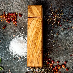 Pode incluir: Um moinho de sal e pimenta de madeira de oliveira. O moinho est&aacute; sobre um fundo cinza com gr&atilde;os de pimenta vermelha, gr&atilde;os de pimenta preta e cristais de sal branco.